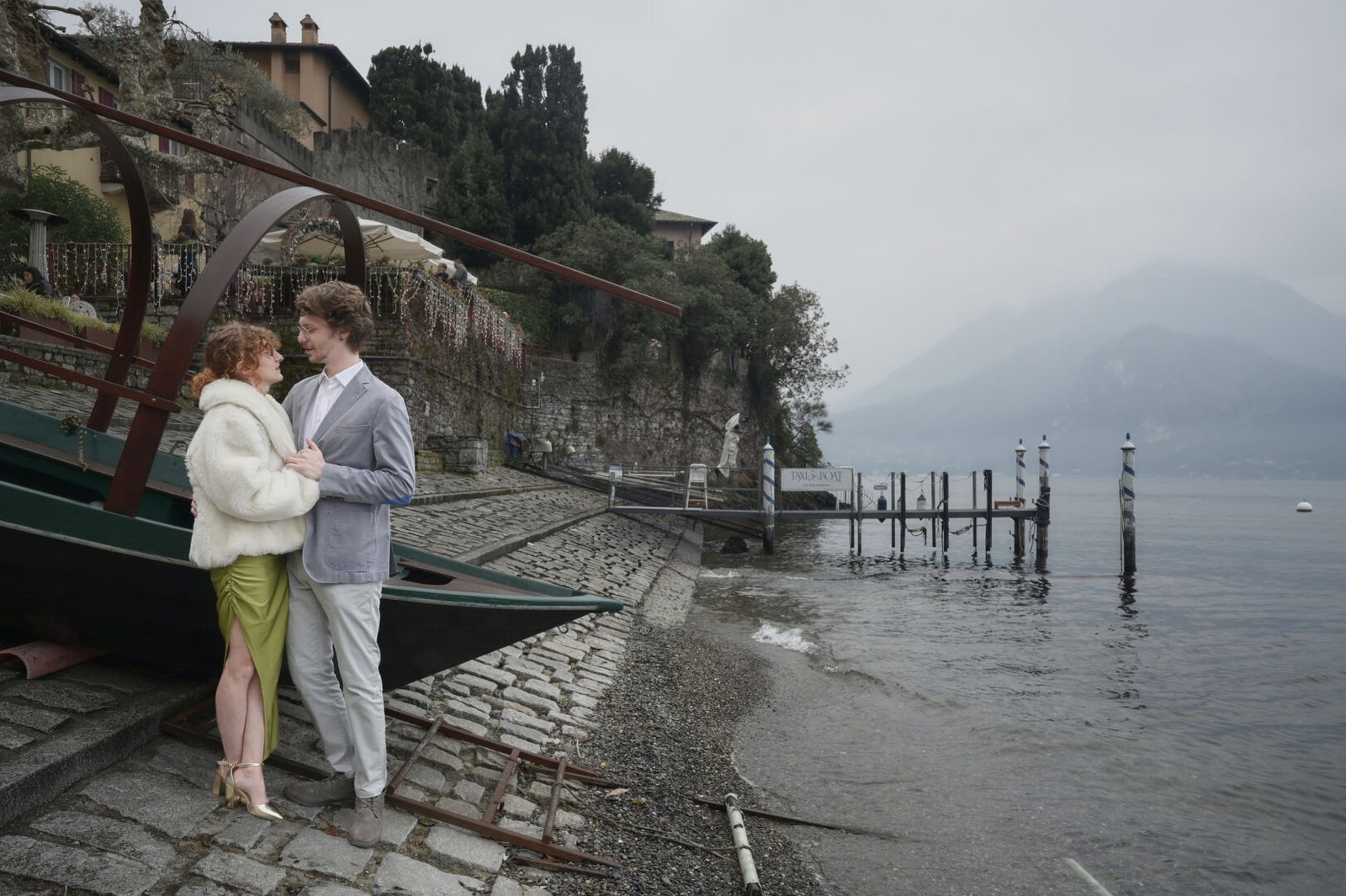 Marco and Giorgia stand close together beside a wooden boat and stone shoreline in Varenna.