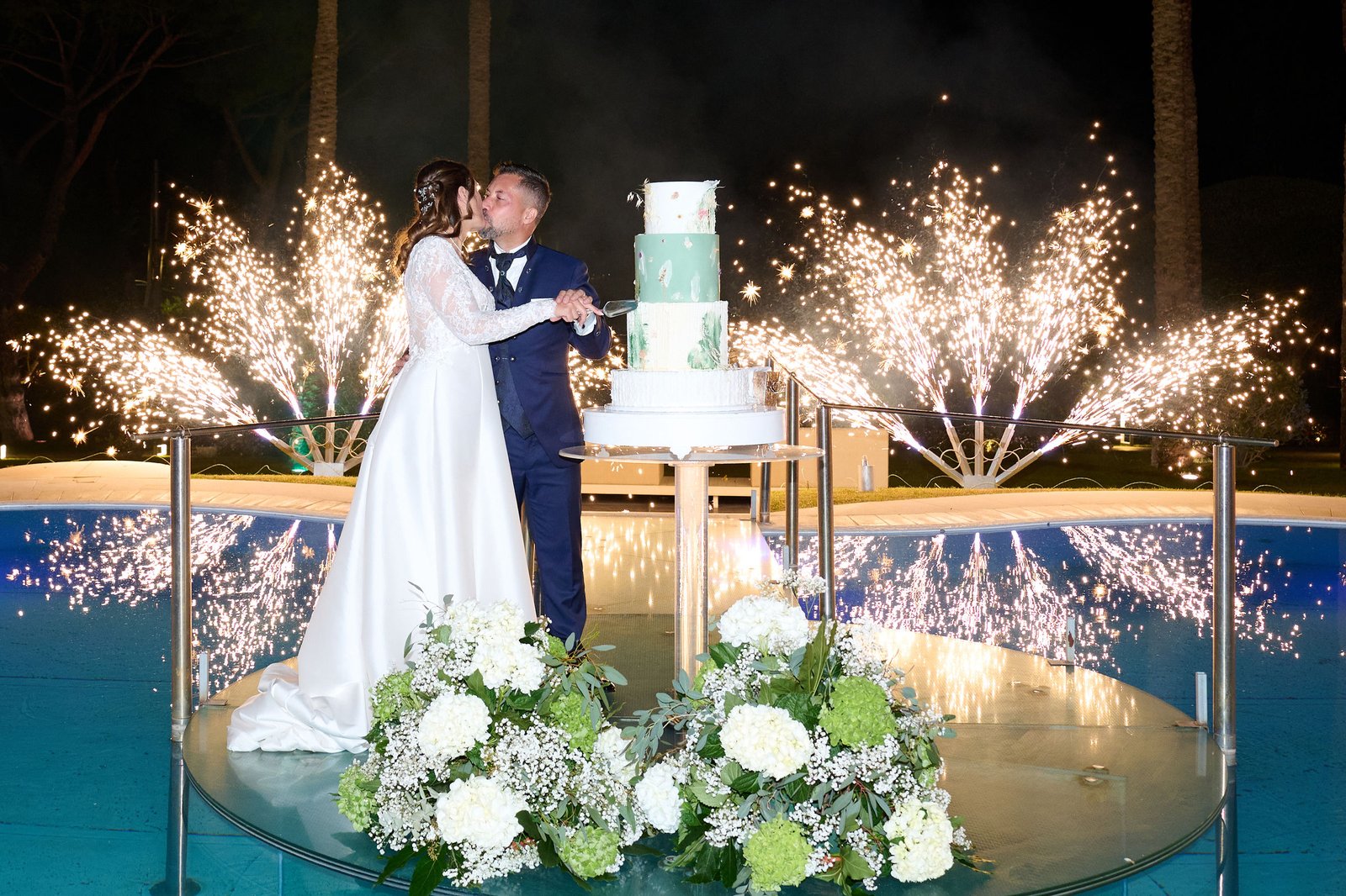 Alessandro and Mariangela kissing beside their wedding cake, surrounded by spark fountains reflected in the pool at Villa Vittoria.