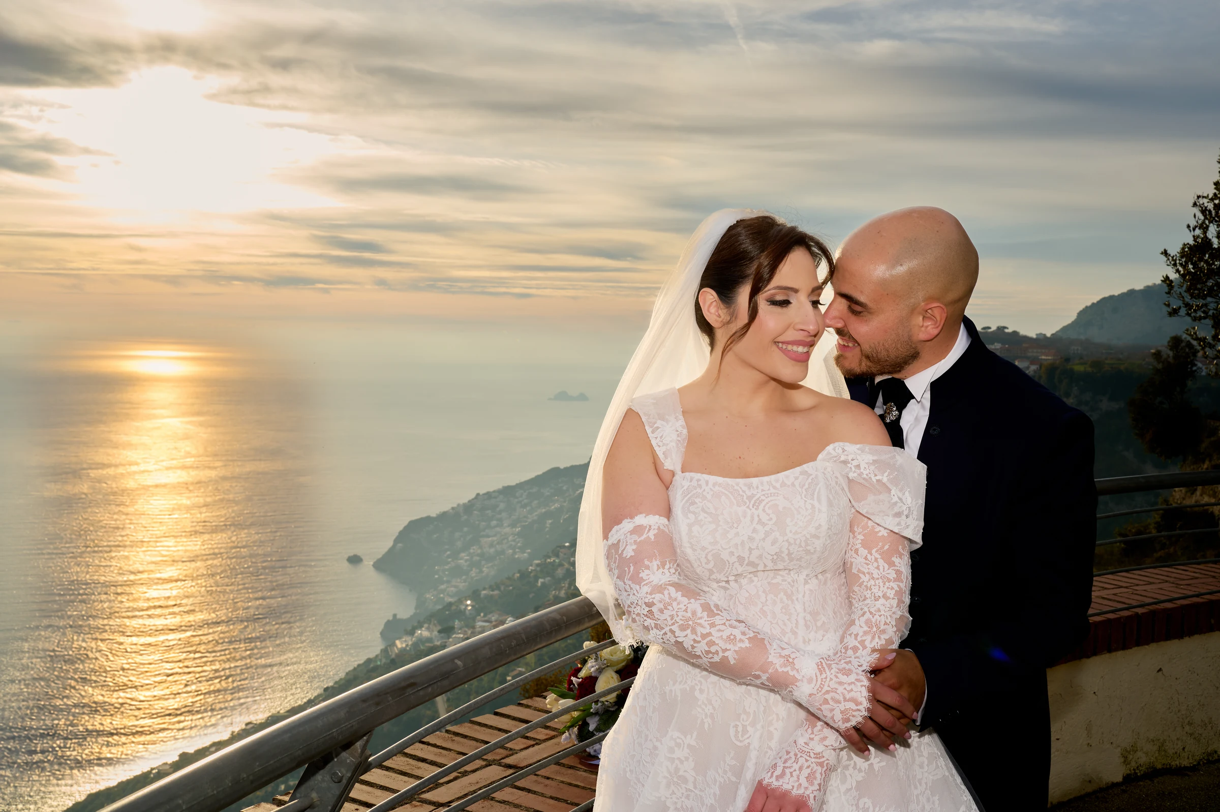 Ciro embracing Rosa at the Belvedere in Agerola, with the Amalfi Coast and sunlight reflected on the sea behind them.