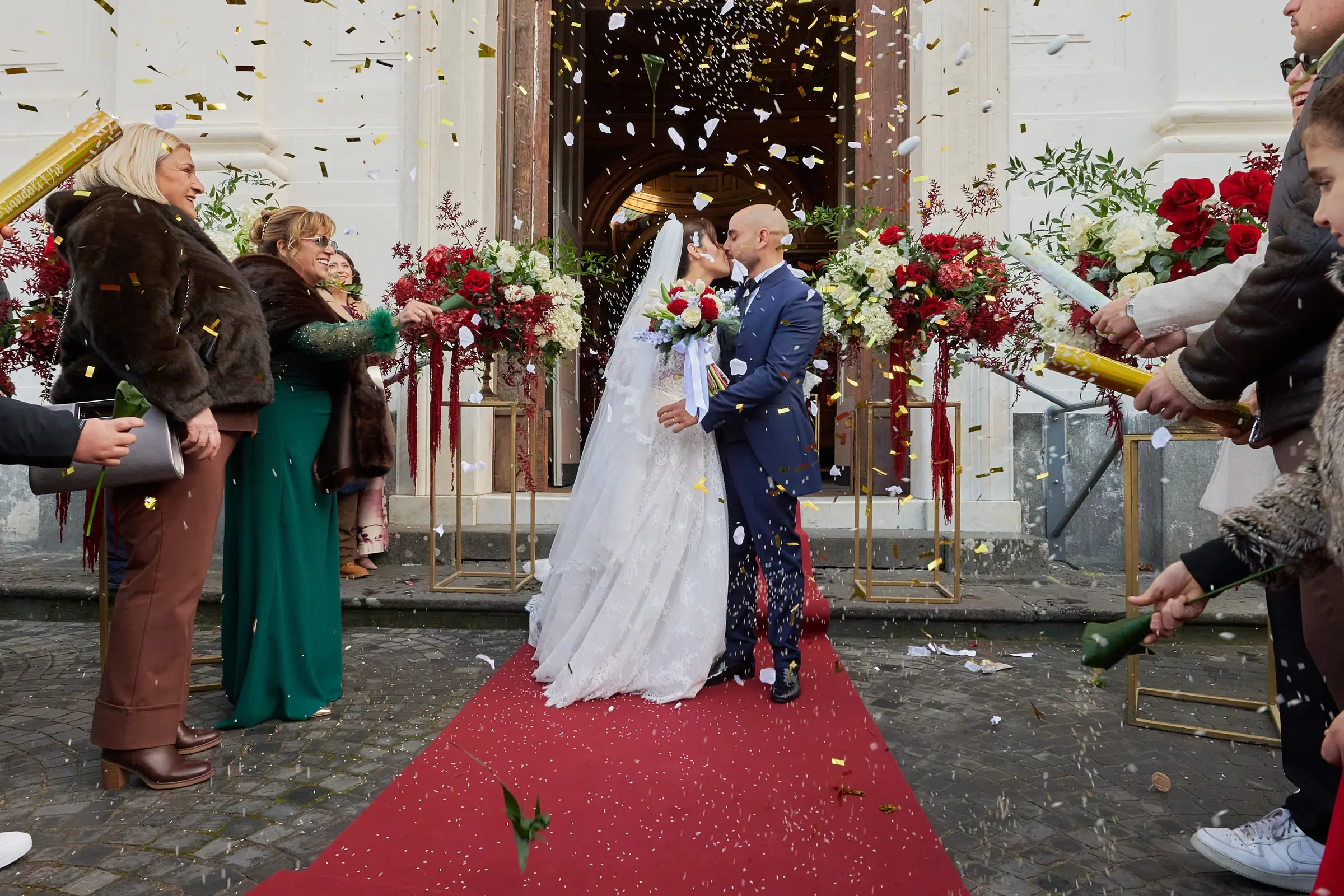 Ciro and Rosa kissing beneath confetti outside the sanctuary in Gragnano, surrounded by guests.
