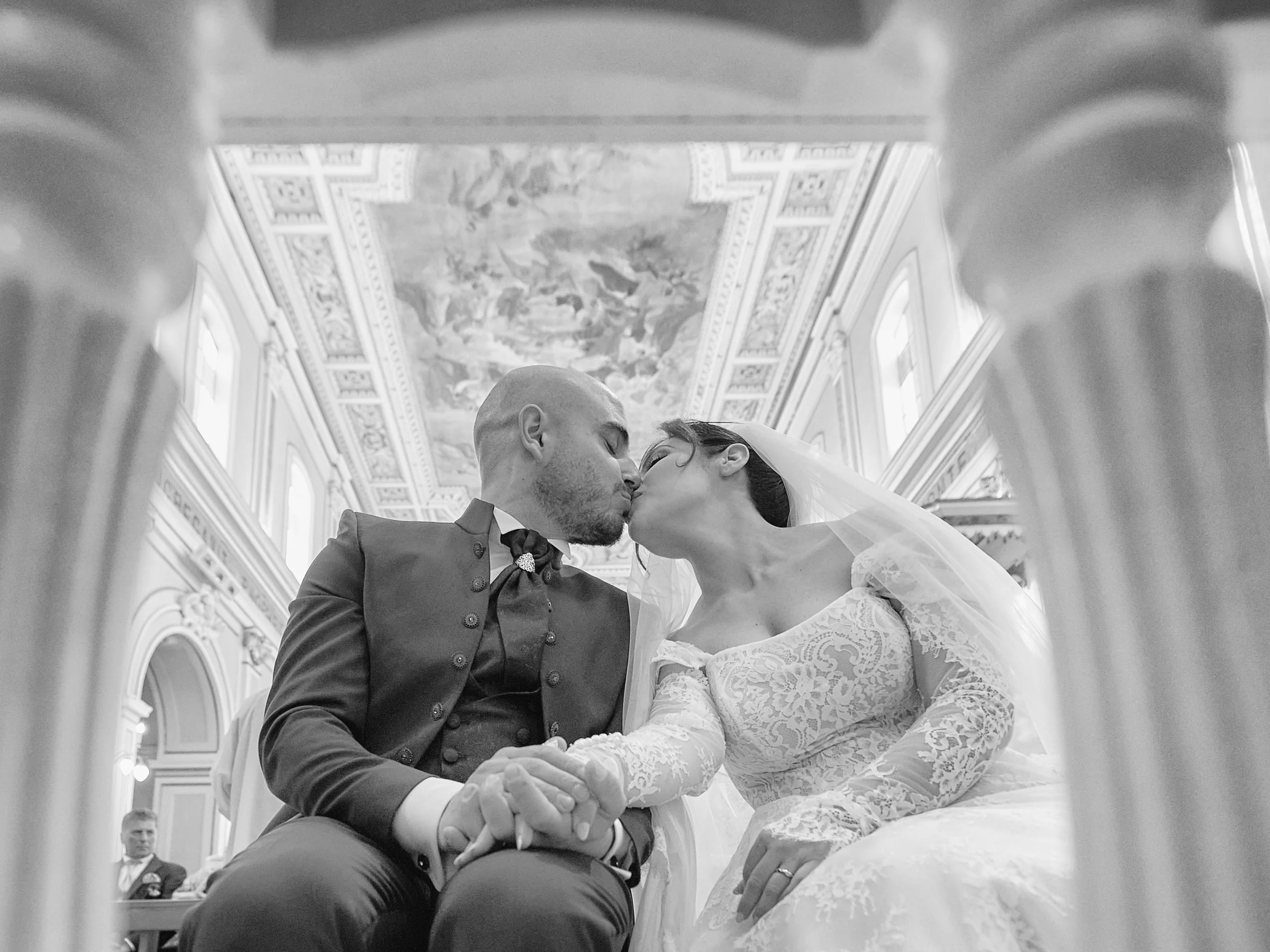 Black and white low-angle photograph of Ciro and Rosa kissing beneath the decorated ceiling of the sanctuary in Gragnano.