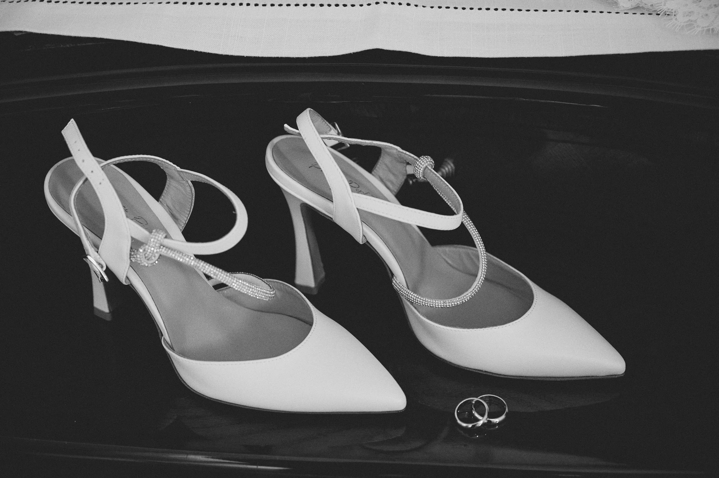 White bridal shoes and wedding rings arranged together, photographed in black and white.