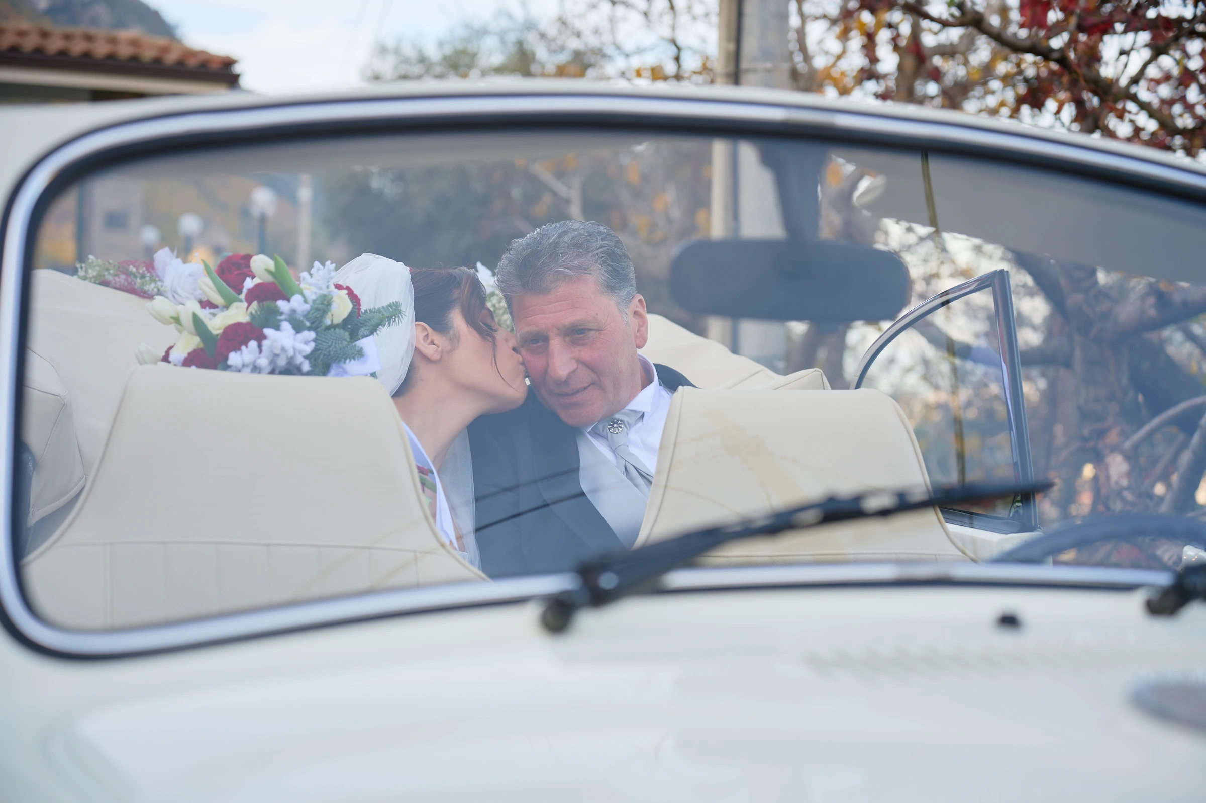 Rosa kissing her father on the cheek inside the wedding car, seen through the windscreen.