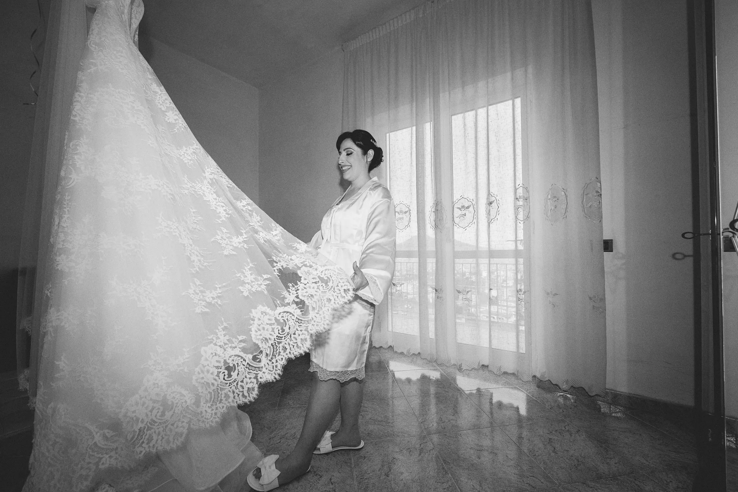 Rosa holding her lace wedding dress beside a window before getting dressed, in black and white.