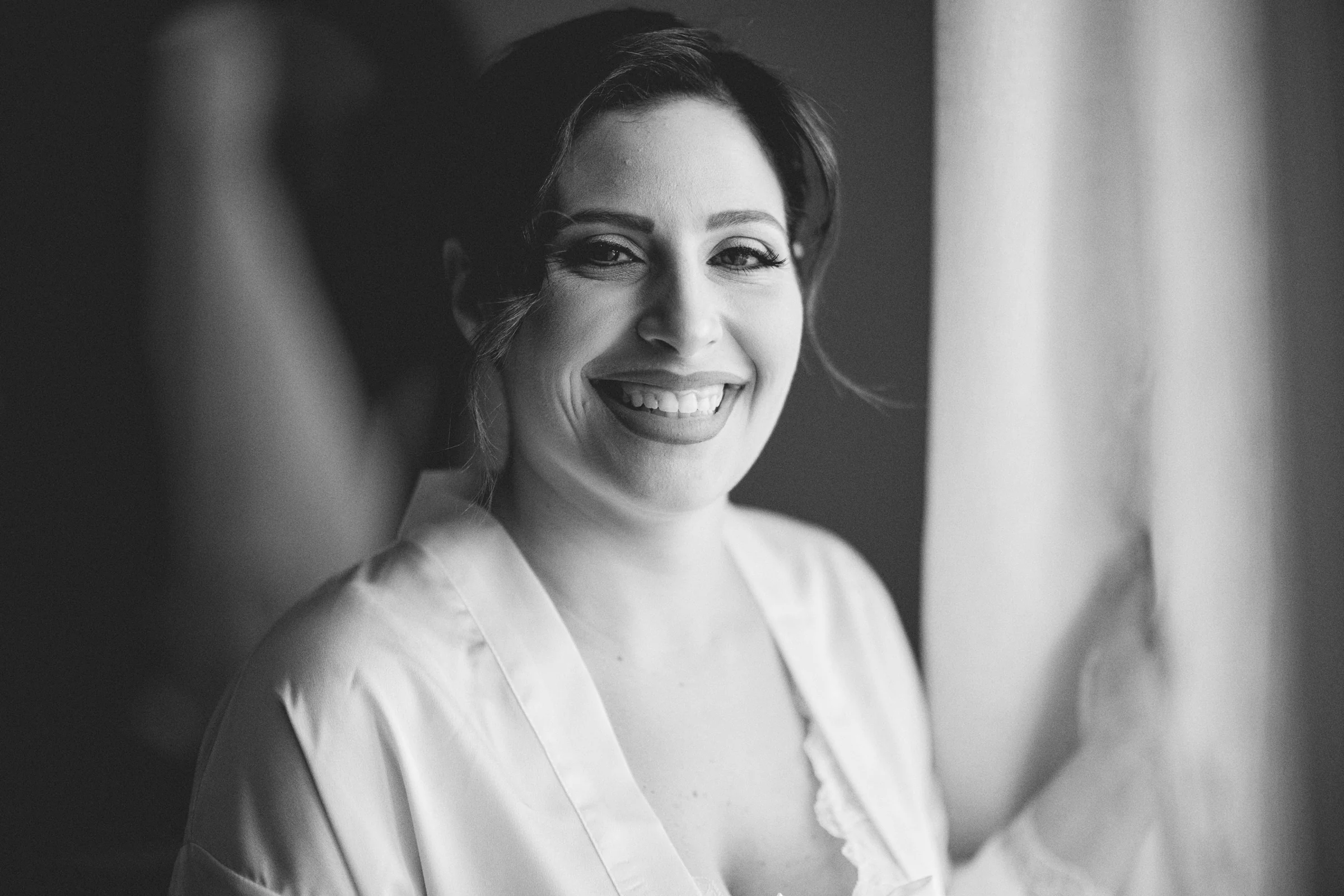 Rosa smiling towards the camera beside a window during her bridal preparations, in black and white.