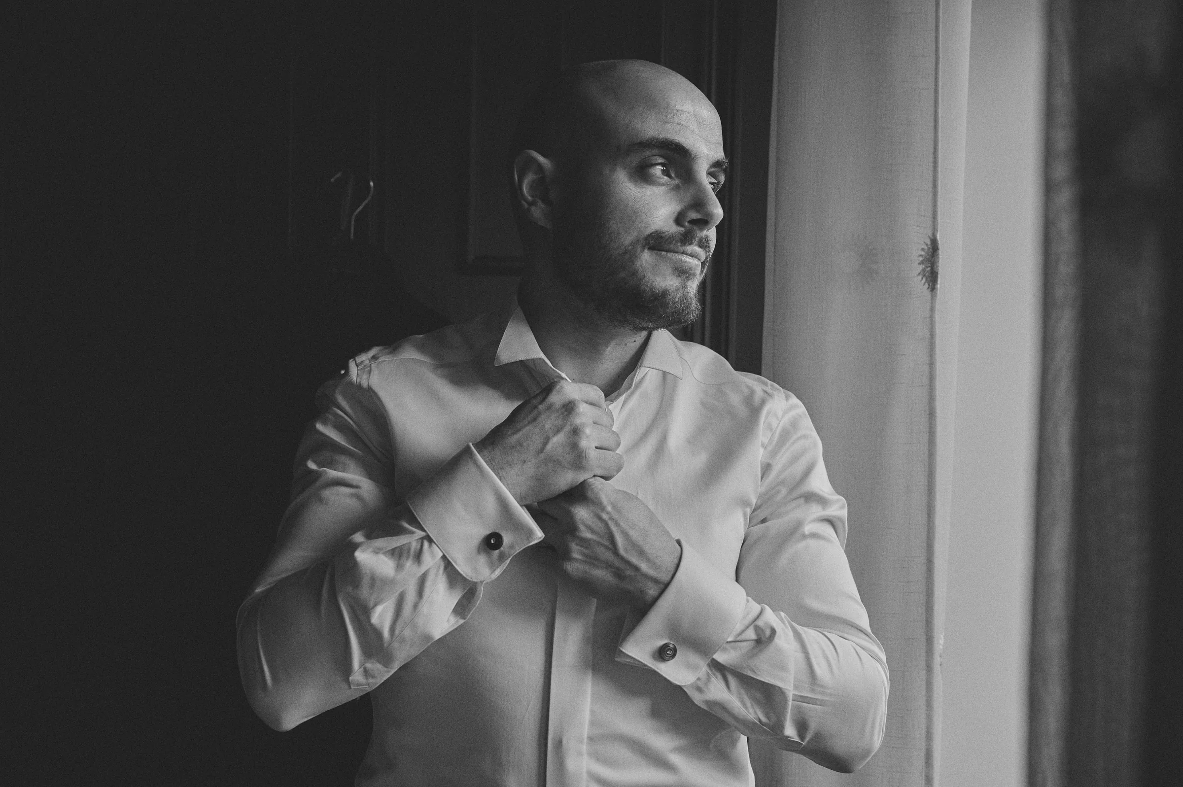 Ciro adjusting his shirt beside a window on his wedding morning, photographed in black and white.