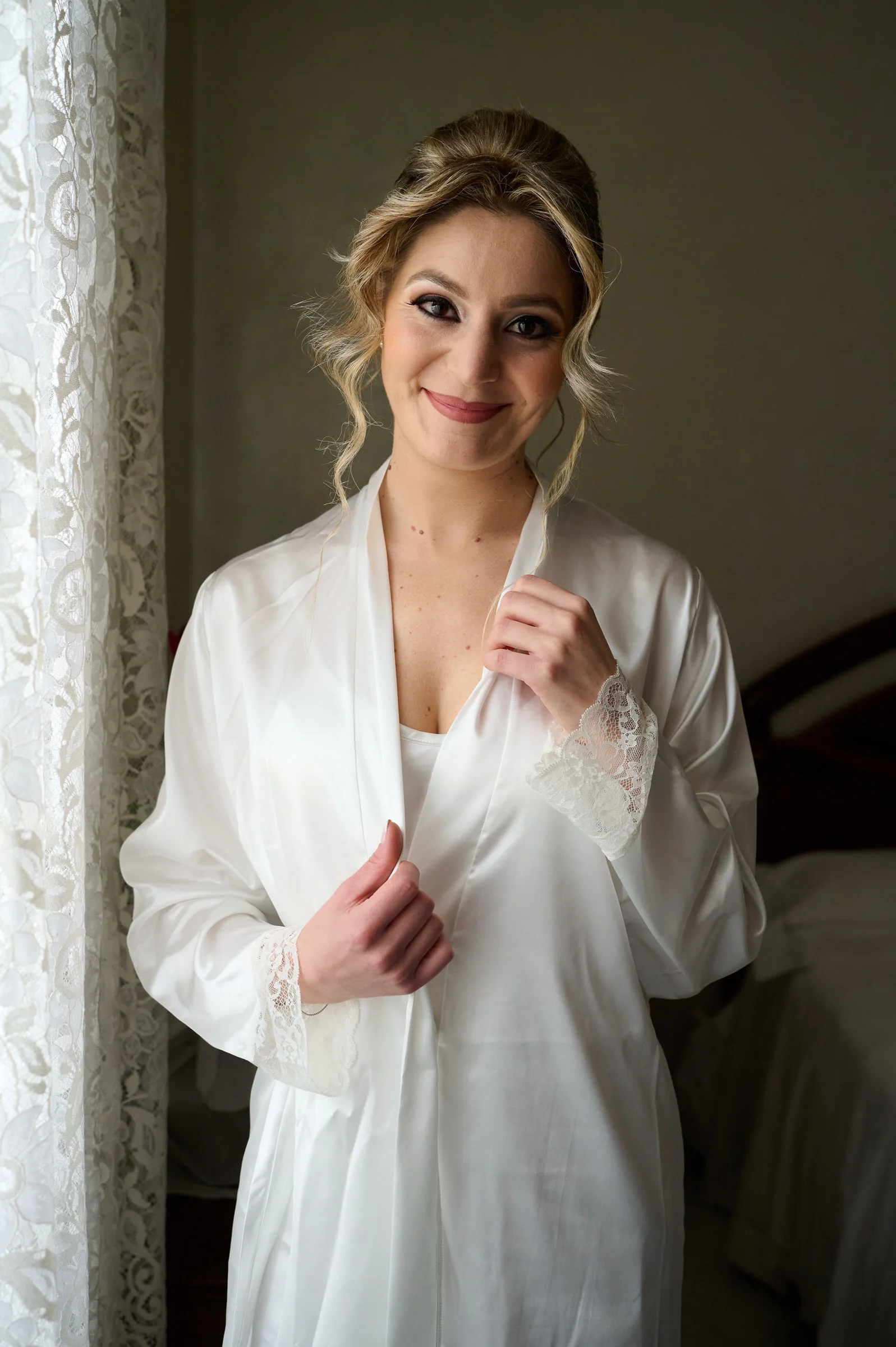Clementina smiling in a bridal robe beside a window during preparations in Gragnano.